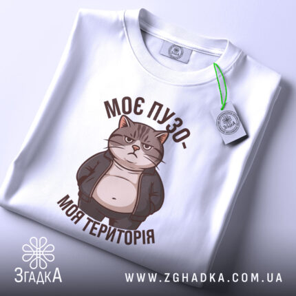 Футболка пузо терапія, складена біла футболка, зображена з верхнього ракурсу, фон світлий, принт з котом у куртці, текст "МОЄ ПУЗО- МОЯ ТЕРИТОРІЯ" в коричневому кольорі, на футболці є етикетка з зеленим шнурком.