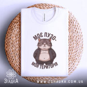 Футболка пузо терапія, складена біла футболка, зображена зверху на плетеній кошику, фон світлий, принт з котом у куртці, текст "МОЄ ПУЗО- МОЯ ТЕРИТОРІЯ" в коричневому кольорі.