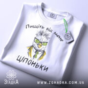 Футболка пищіть від мене. Біла футболка, складена на поверхні, з видимим принтом птаха в окулярах та жовтому піджаку. На футболці текст "Ціпоньки". Зелена етикетка прикріплена до коміра. Фон світлий, однорідний.