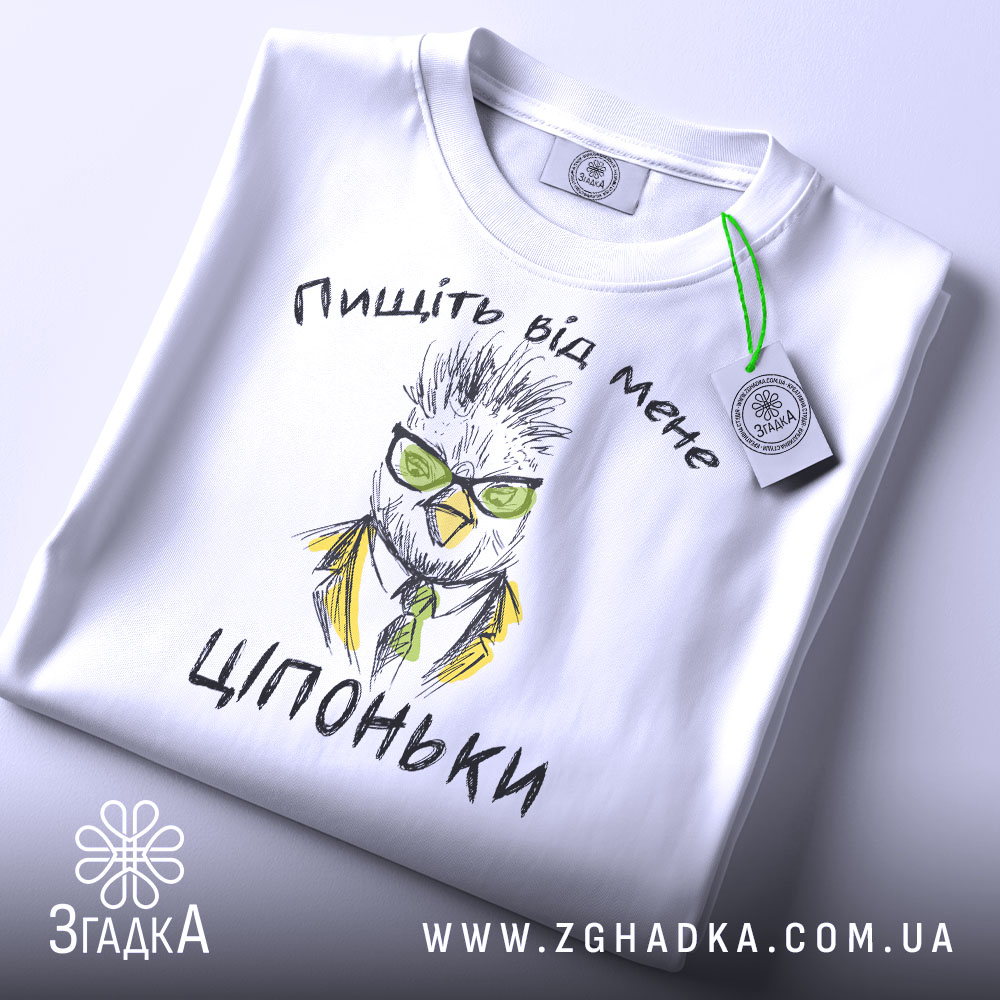 Футболка пищіть від мене. Біла футболка, складена на поверхні, з видимим принтом птаха в окулярах та жовтому піджаку. На футболці текст "Ціпоньки". Зелена етикетка прикріплена до коміра. Фон світлий, однорідний.