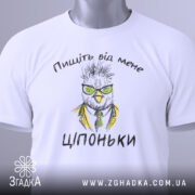 Футболка пищіть від мене. Біла футболка, складена на поверхні, з видимим принтом птаха в окулярах та жовтому піджаку. На футболці текст "Ціпоньки". Зелена етикетка прикріплена до коміра. Фон світлий, однорідний.