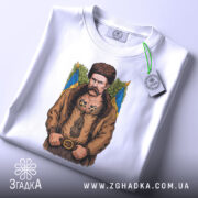 Футболка письменник Шевченко, біла, зображена на столі, фон світло-сірий, принт з зображенням чоловіка в традиційному одязі, на футболці є етикетка з зеленим шнурком.