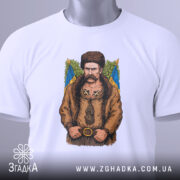 Футболка письменник Шевченко, біла, зображена на столі, фон світло-сірий, принт з зображенням чоловіка в традиційному одязі, на футболці є етикетка з зеленим шнурком.