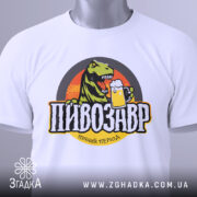 Футболка пивозавр пінний період, біла, зображена зверху, складена, фон світлий, принт з динозавром, що тримає пиво, кольори яскраві, на футболці є етикетка з зеленим шнурком.