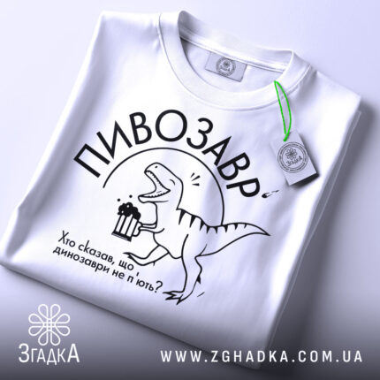 Футболка пивозавр білого кольору, складена на поверхні, фон світлий, принт з динозавром, що тримає пиво, текст: "Хто сказав, що динозаври не п'ють?", на футболці є етикетка з зеленим шнурком.