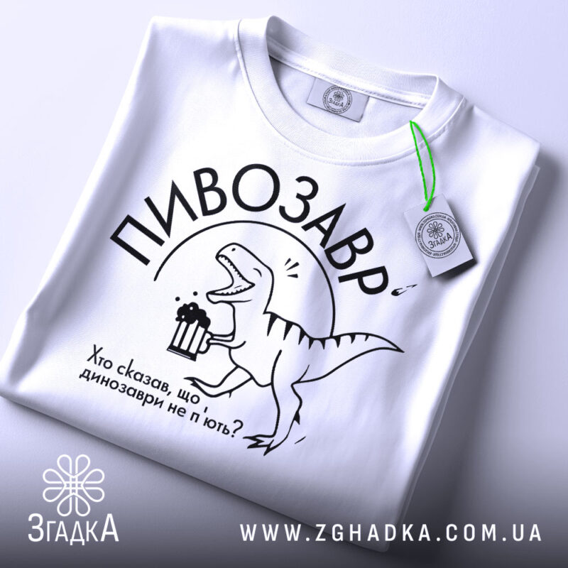 Футболка пивозавр білого кольору, складена на поверхні, фон світлий, принт з динозавром, що тримає пиво, текст: "Хто сказав, що динозаври не п'ють?", на футболці є етикетка з зеленим шнурком.