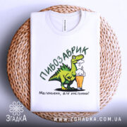 Футболка пивозаврик білого кольору, складена на плетеній основі, фон світлий, принт з динозавром, що тримає пиво, текст: 'Маленький, але хмільний!'.