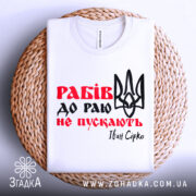 Футболка рабів не пускають, біла футболка складена на плетеній кошику, фон світлий, принт з червоним текстом "Рабів до раю не пускають" та чорним тризубом, видна етикетка на комірці.