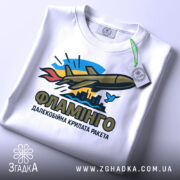 Футболка ракета Фламінго білого кольору, складена на столі, фон світло-сірий, принт з ракетою, містом та птахом, прикріплена етикетка з зеленим шнурком.