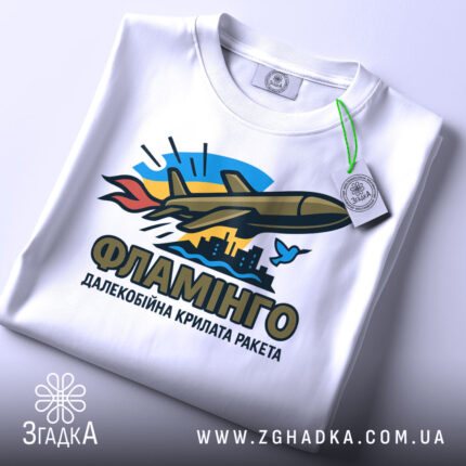 Футболка ракета Фламінго білого кольору, складена на столі, фон світло-сірий, принт з ракетою, містом та птахом, прикріплена етикетка з зеленим шнурком.