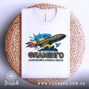 Футболка ракета Фламінго білого кольору, складена на плетеному кошику, фон світлий, принт з ракетою, містом та птахом, видно округлі краї горловини.