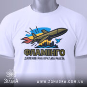 Футболка ракета Фламінго білого кольору, складена на столі, фон світло-сірий, принт з ракетою, містом та птахом, прикріплена етикетка з зеленим шнурком.