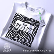 Футболка реаліст білого кольору, складена на поверхні, з видимим принтом чорного кольору з абстрактними лініями та написом 'THE REALIST'. На комірці є етикетка з зеленим шнурком.