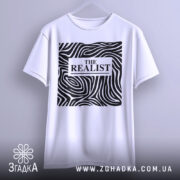 Футболка реаліст білого кольору, підвішена на вішалці, фон світло-сірий. Принт чорного кольору з абстрактними лініями та написом 'THE REALIST' у центрі.