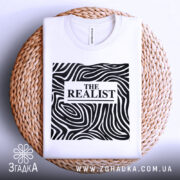 Футболка реаліст білого кольору, складена на плетеній кошику, фон світло-сірий. Принт чорного кольору з абстрактними лініями та написом 'THE REALIST' у центрі.