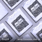 Футболка реаліст білого кольору, зображена на манекені з правого ракурсу, фон світло-сірий. Принт чорного кольору з абстрактними лініями та написом 'THE REALIST' у центрі.