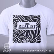 Футболка реаліст білого кольору, складена на поверхні, з видимим принтом чорного кольору з абстрактними лініями та написом 'THE REALIST'. На комірці є етикетка з зеленим шнурком.