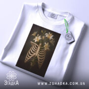 Футболка ребра розквітли, біла футболка з принтом, що зображує скелетні ребра з ліліями, розкладена на поверхні, з етикеткою, на світлому фоні, з легким нахилом.