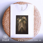 Футболка ребра розквітли, біла футболка з принтом, що зображує скелетні ребра з ліліями, розкладена на плетеній основі, на світлому фоні, з легким нахилом.