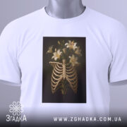 Футболка ребра розквітли, біла футболка з принтом, що зображує скелетні ребра з ліліями, розкладена на поверхні, з етикеткою, на світлому фоні, з легким нахилом.