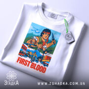 Футболка Рембо перша кров білого кольору, зображена з верхнього ракурсу, на ній підвішена етикетка, фон світло-сірий, принт яскравий, з персонажами та написом 'FIRST BLOOD'.