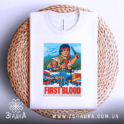 Футболка Рембо перша кров білого кольору, зображена на плетеній основі з верхнього ракурсу, фон білий, принт яскравий, з персонажами та написом 'FIRST BLOOD'.