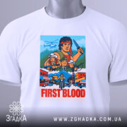 Футболка Рембо перша кров білого кольору, зображена з верхнього ракурсу, на ній підвішена етикетка, фон світло-сірий, принт яскравий, з персонажами та написом 'FIRST BLOOD'.