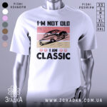 Футболка ретро авто білого кольору, зображена з фронтального ракурсу на манекені, фон світло-сірий, принт з автомобілем у стилі ретро на рожевому та бежевому фоні, текст "I'M NOT OLD I AM CLASSIC" великими чорними літерами.