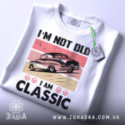 Футболка ретро авто білого кольору, зображена зверху, на ній прикріплена етикетка, фон світло-сірий, принт з автомобілем у стилі ретро на рожевому та бежевому фоні, текст "I'M NOT OLD I AM CLASSIC" великими чорними літерами.