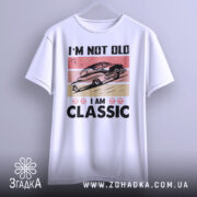 Футболка ретро авто білого кольору, зображена з фронтального ракурсу на вішалці, фон світло-сірий, принт з автомобілем у стилі ретро на рожевому та бежевому фоні, текст "I'M NOT OLD I AM CLASSIC" великими чорними літерами.