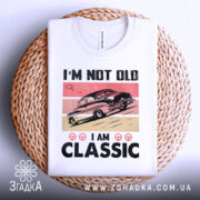 Футболка ретро авто білого кольору, зображена зверху на плетеній кошику, фон світло-сірий, принт з автомобілем у стилі ретро на рожевому та бежевому фоні, текст "I'M NOT OLD I AM CLASSIC" великими чорними літерами.