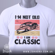 Футболка ретро авто білого кольору, зображена з фронтального ракурсу на манекені, фон світло-сірий, принт з автомобілем у стилі ретро на рожевому та бежевому фоні, текст "I'M NOT OLD I AM CLASSIC" великими чорними літерами.