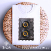 Футболка ретро касета білого кольору, зображена на плетеній основі, ракурс зверху, фон світло-сірий, принт з касетою з жовтими котушками, футболка акуратно складена.