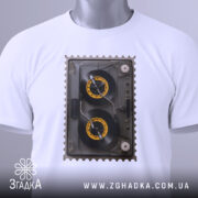 Футболка ретро касета білого кольору, зображена на столі, ракурс зверху, фон світло-сірий, принт з касетою з жовтими котушками, на футболці є етикетка з логотипом.