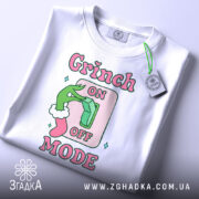 Футболка режим Грінча, біла, зображена зверху, фон світлий, принт з зеленим рукою Грінча, що натискає на вимикач, текст 'ON OFF MODE' в рожевих і зелених кольорах, з біркою на шнурку.