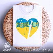 Футболка рідний дім, біла, складена на плетеному кошику, фон світло-сірий, принт у формі серця з жовто-блакитними кольорами та написом 'РІДНИЙ ДІМ', зображення меча в центрі.