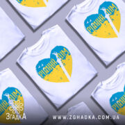 Футболка рідний дім, біла, зображена на манекені, фон світло-сірий, принт у формі серця з жовто-блакитними кольорами та написом 'РІДНИЙ ДІМ' у верхній частині, зображення меча в центрі.