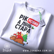 Футболка рік новий, біла, складена на столі, фон світло-сірий, принт з оленем у червоному капелюсі, ялинкою та подарунками, текст "РІК новий А ЛІНЬ СТАРА", з етикеткою на комірці.
