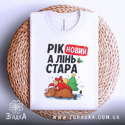 Футболка рік новий, біла, складена на плетеній кошику, фон білий, принт з оленем у червоному капелюсі, ялинкою та подарунками, текст "РІК новий А ЛІНЬ СТАРА".
