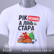 Футболка рік новий, біла, складена на столі, фон світло-сірий, принт з оленем у червоному капелюсі, ялинкою та подарунками, текст "РІК новий А ЛІНЬ СТАРА", з етикеткою на комірці.