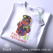 Футболка рівень 40 пройдено, біла футболка на столі, ракурс зверху, фон світло-сірий, принт з персонажем у капелюсі та окулярах, який тримає геймпад, текст 'РІВЕНЬ 40 пройдено' внизу, на футболці є етикетка.