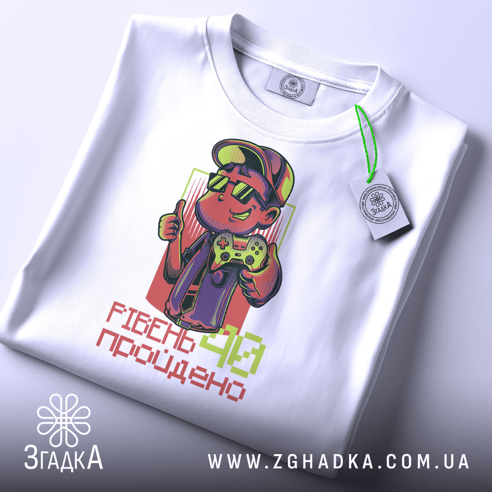 Футболка рівень 40 пройдено, біла футболка на столі, ракурс зверху, фон світло-сірий, принт з персонажем у капелюсі та окулярах, який тримає геймпад, текст 'РІВЕНЬ 40 пройдено' внизу, на футболці є етикетка.
