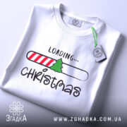 Футболка різдвяний прогрес, біла, складена на поверхні, зображена зверху, фон світлий, принт містить напис 'LOADING...' та малюнок ялинки на смужці з червоними і білими смугами, на футболці є етикетка.