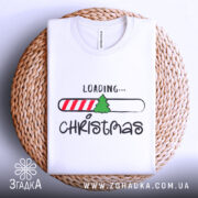Футболка різдвяний прогрес, біла, складена на круглій кошику, фон світлий, принт містить напис 'LOADING...' та малюнок ялинки на смужці з червоними і білими смугами.