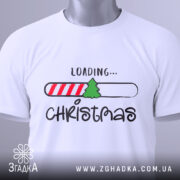 Футболка різдвяний прогрес, біла, складена на поверхні, зображена зверху, фон світлий, принт містить напис 'LOADING...' та малюнок ялинки на смужці з червоними і білими смугами, на футболці є етикетка.