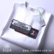Футболка робот автономний, біла, складена на столі, фон світлий, принт чорного кольору з червоними елементами, текст на українській мові, на футболці є етикетка.