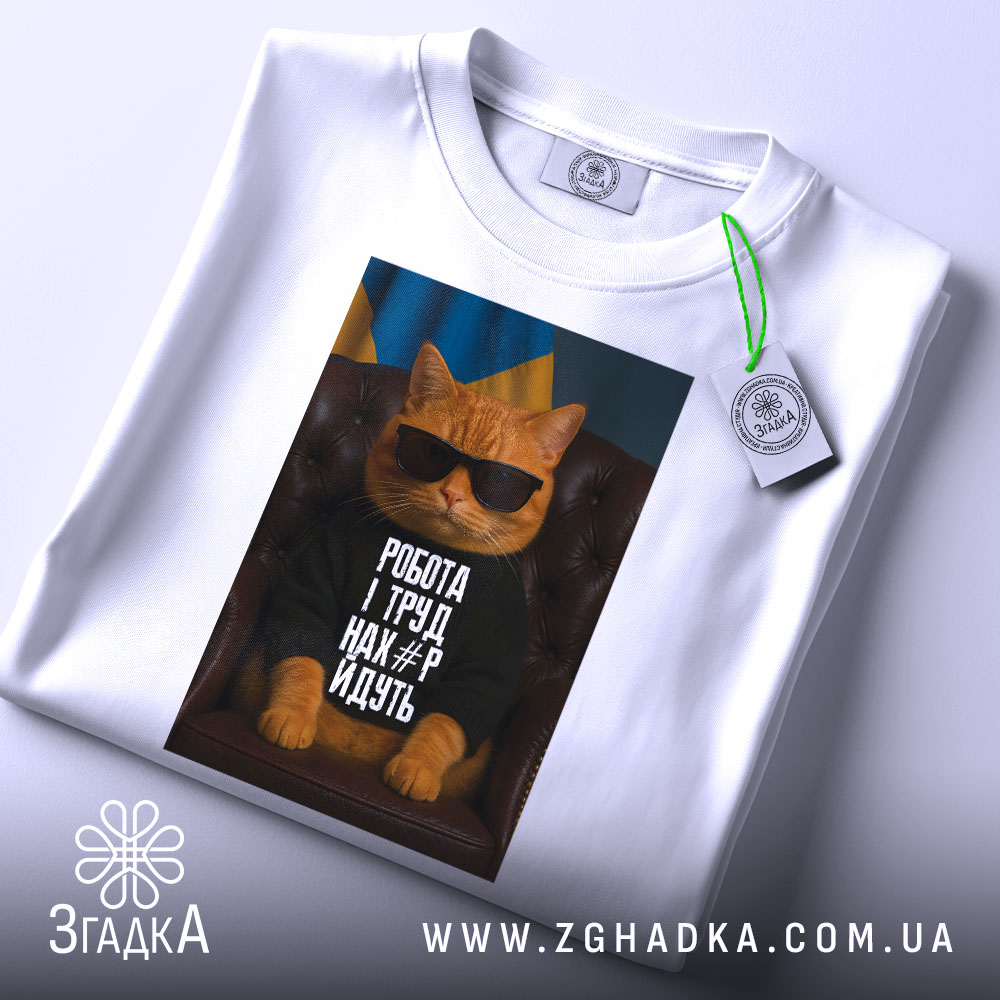 Футболка робота і труд. Біла футболка, складена на поверхні, з принтом кота в окулярах. На фоні видно український прапор, а також етикетка з зеленим шнурком.