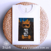 Футболка робота і труд. Біла футболка, складена на плетеній кошику, з принтом кота в окулярах. Фон нейтральний, що підкреслює кольори футболки та принта.