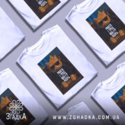 Футболка робота і труд. Біла футболка з принтом, на якому зображено кота в окулярах, сидячого на кріслі. Фон складається з українського прапора, що підкреслює кольори принта.