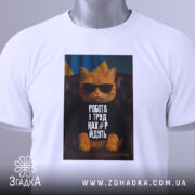 Футболка робота і труд. Біла футболка, складена на поверхні, з принтом кота в окулярах. На фоні видно український прапор, а також етикетка з зеленим шнурком.